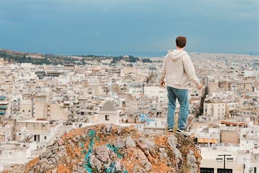 Das Bild zeigt einen Mann, den man von hinten sieht. Der Mann steht auf einem Felsen über einer Stadt und schaut Richtung Stadt.